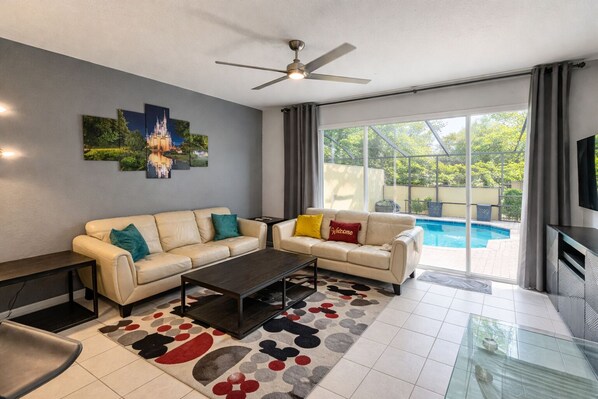 A modern living room with a beige sofa, a coffee table, and a flat-screen TV. There is a ceiling fan, a large window with curtains, and a pool visible through the glass doors. - Disney Villa| Private Pool|5 mins to Disney|Resort amenities (Kissimmee)