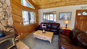 A cozy cabin interior with a stone wall, wooden ceiling, and large windows. There is a leather sofa, a wooden coffee table, and a fireplace. - Preferred Parking Pass to Dodge Ridge (Cold Springs)