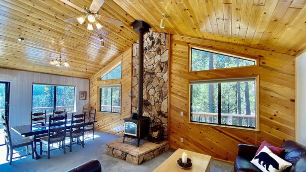 A cozy cabin interior with a stone fireplace, wooden walls, large windows, a dining area, and a leather sofa. - Preferred Parking Pass to Dodge Ridge (Cold Springs)