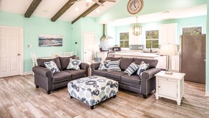 A spacious living room with two sofas and a matching ottoman, a ceiling fan, and a kitchen area in the background.