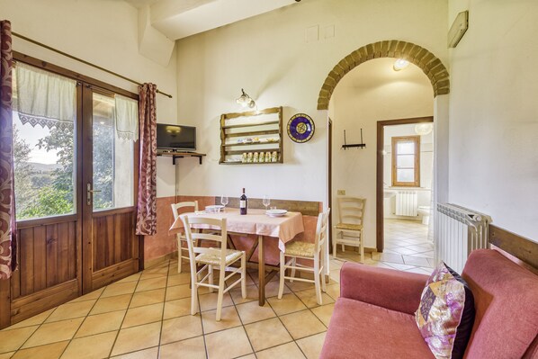 Uma área de refeições com uma mesa posta para dois, uma televisão na parede e vista para o jardim pela janela.