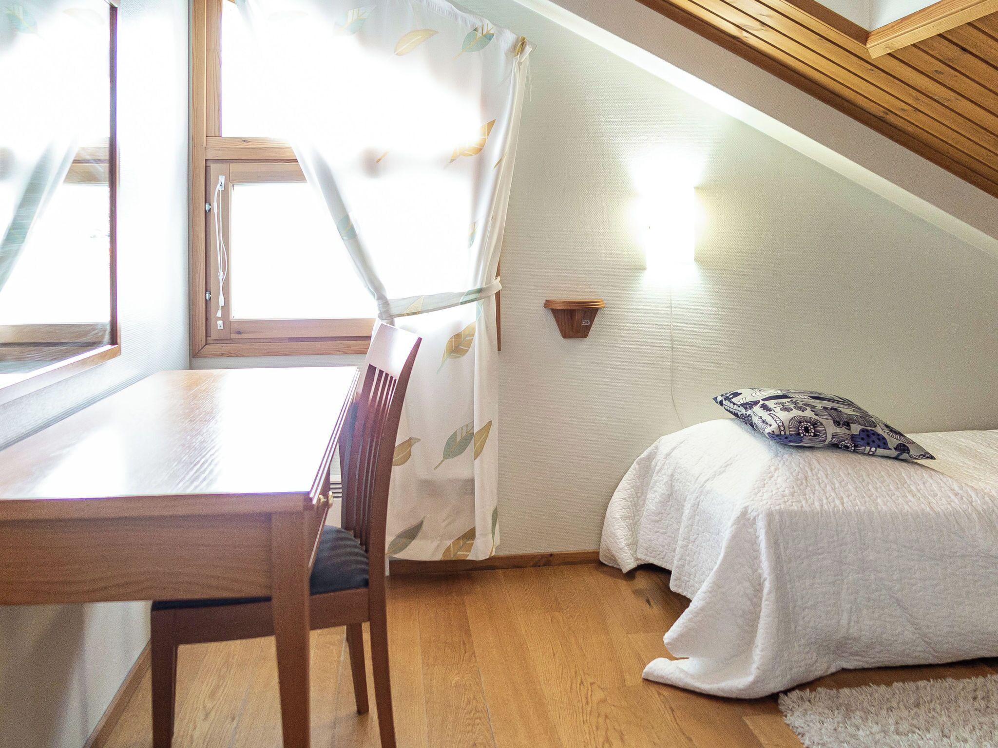 A bedroom with a wooden desk, a chair, a bed with a patterned pillow, and a wall-mounted light fixture.
