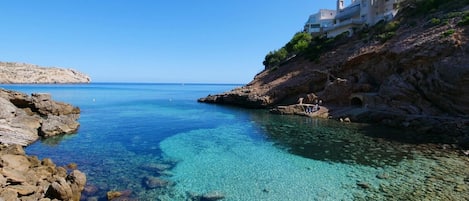 A coastal scene with clear blue water, rocky shore, and a building on a cliff.