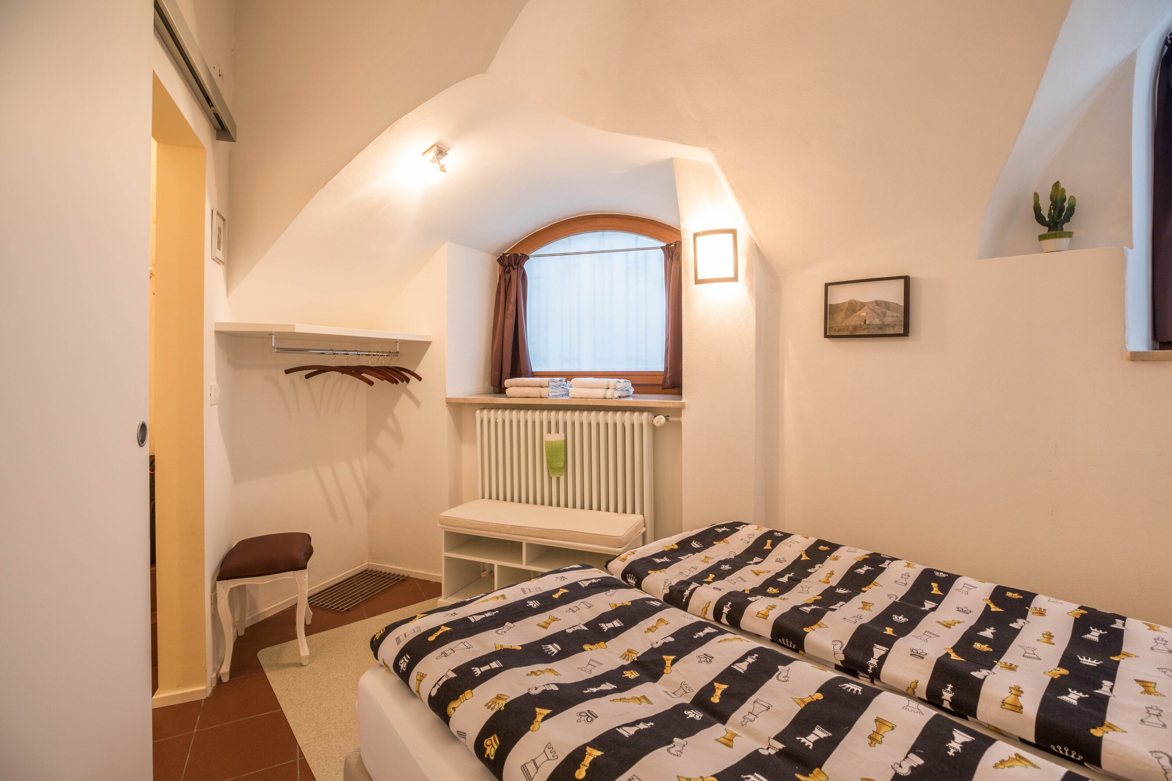Ein Schlafzimmer mit Bett, Fenster, Heizkörper, einer Bank und einem Wandregal.
