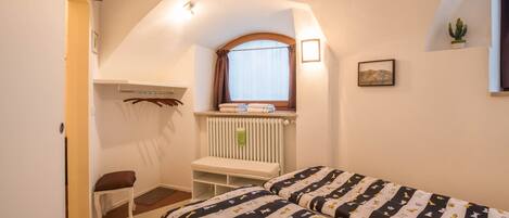 Ein Schlafzimmer mit Bett, Fenster, Heizkörper, einer Bank und einem Wandregal.