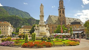 Ein Platz mit einem zentralen Denkmal, Bänken und Blumenbeeten.