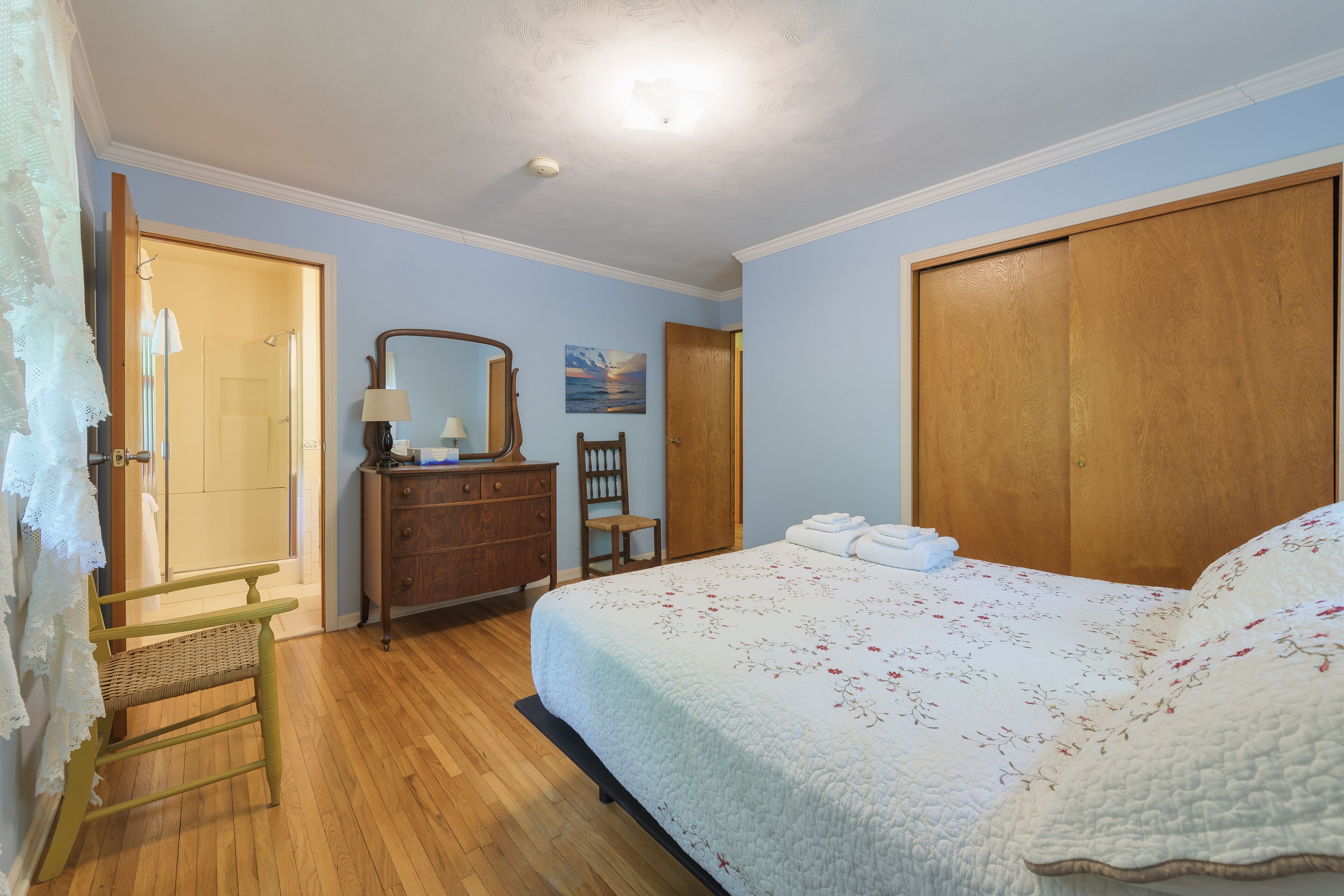 A bedroom with a bed, wooden wardrobe, dresser, chair, and a painting on the wall.