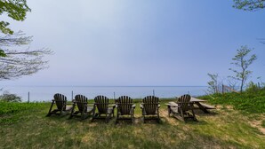 Eine Reihe aus Holz gefertigter Adirondack-Stühle, die auf ein Gewässer ausgerichtet sind, mit einer Rasenfläche und einem Zaun im Hintergrund.
