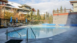 A swimming pool with lounge chairs and umbrellas, surrounded by buildings with a stone wall and a wooden deck. - Mountain View Penthouse 2Bdrm Loft Suite w Pool and 3 Hot Tubs (Canmore)