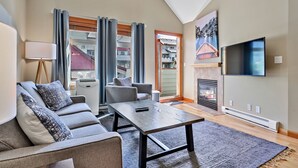 A modern living room with a grey sofa, a coffee table, a flat-screen TV, and a fireplace. - Mountain View Penthouse 2Bdrm Loft Suite w Pool and 3 Hot Tubs (Canmore)