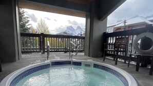 A hot tub with a tiled edge, a metal ladder, and a view of mountains through a wooden railing. - Mountain View Penthouse 2Bdrm Loft Suite w Pool and 3 Hot Tubs (Canmore)