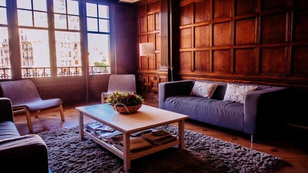 A living room with a wooden paneled wall, a sofa, a chair, a coffee table with a plant, and a window with a view of buildings. - Zentric Hostel (León)