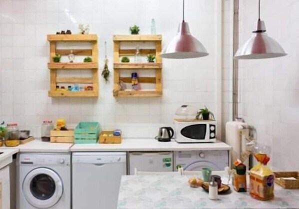 A compact kitchen with a washing machine, microwave, wooden shelves, and pendant lights. - Zentric Hostel (León)