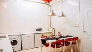 A compact kitchen with a washing machine, a microwave, and a dining table with red chairs. - Zentric Hostel (León)
