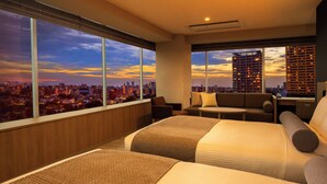 Une chambre d’hôtel avec deux lits, un canapé, une petite table et une vue sur le paysage urbain au coucher du soleil.