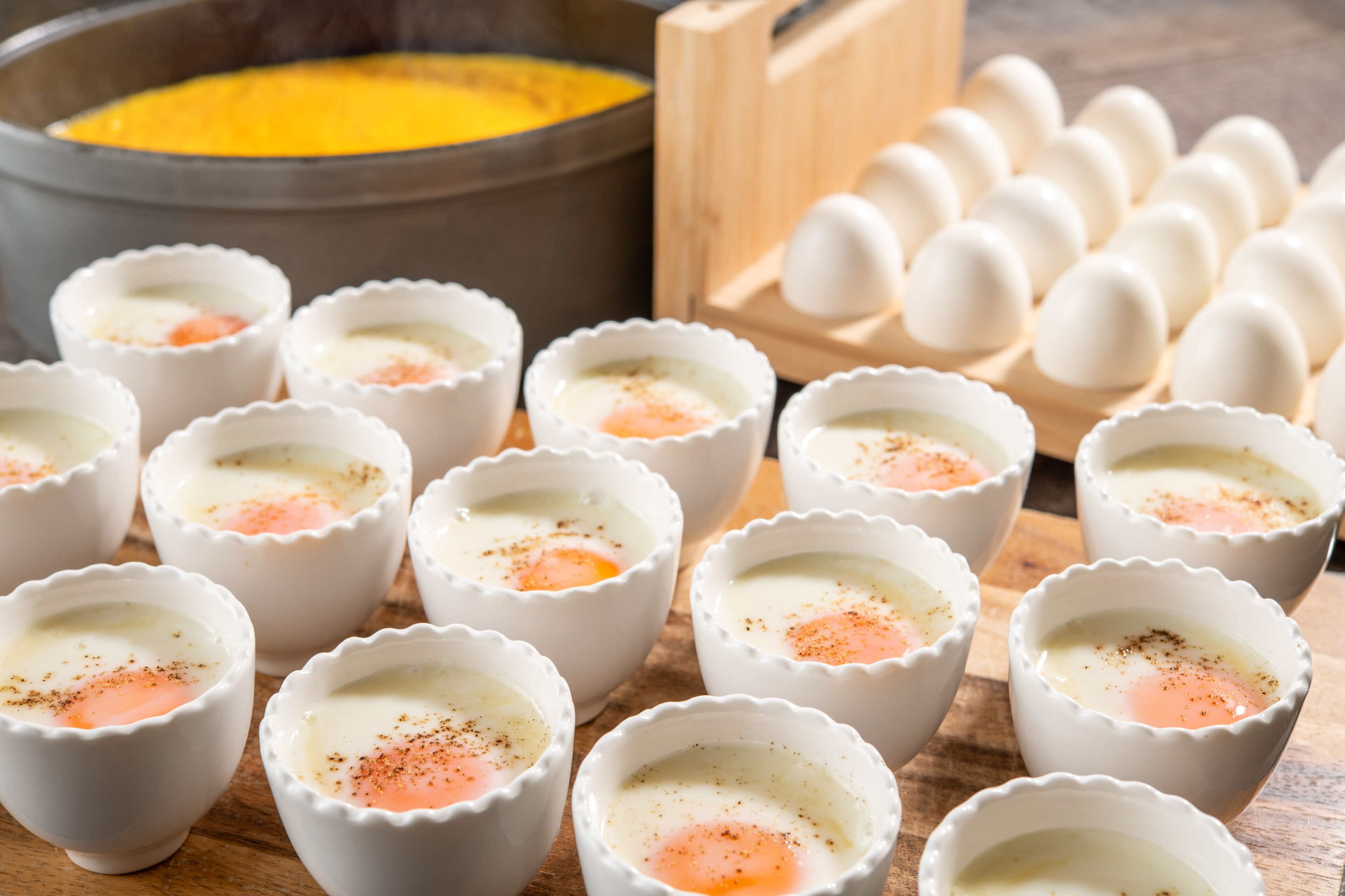 A series of white ramekins with a cooked egg in each, a wooden cutting board, a bowl of yellow liquid, and a carton of eggs.