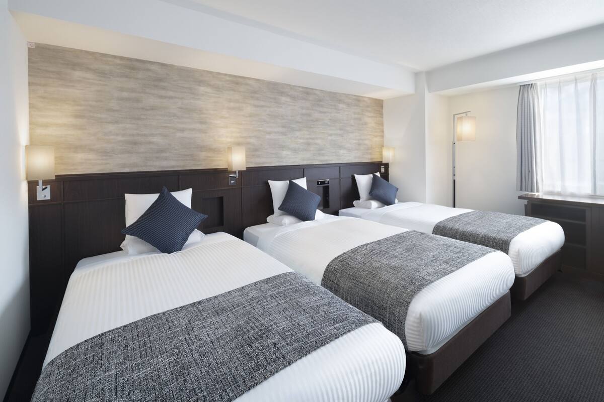 Une chambre d’hôtel avec trois lits, chacun doté de draps blancs et de plaids gris. Des lampes murales sont fixées de chaque côté des lits, et une grande fenêtre est agrémentée de rideaux.