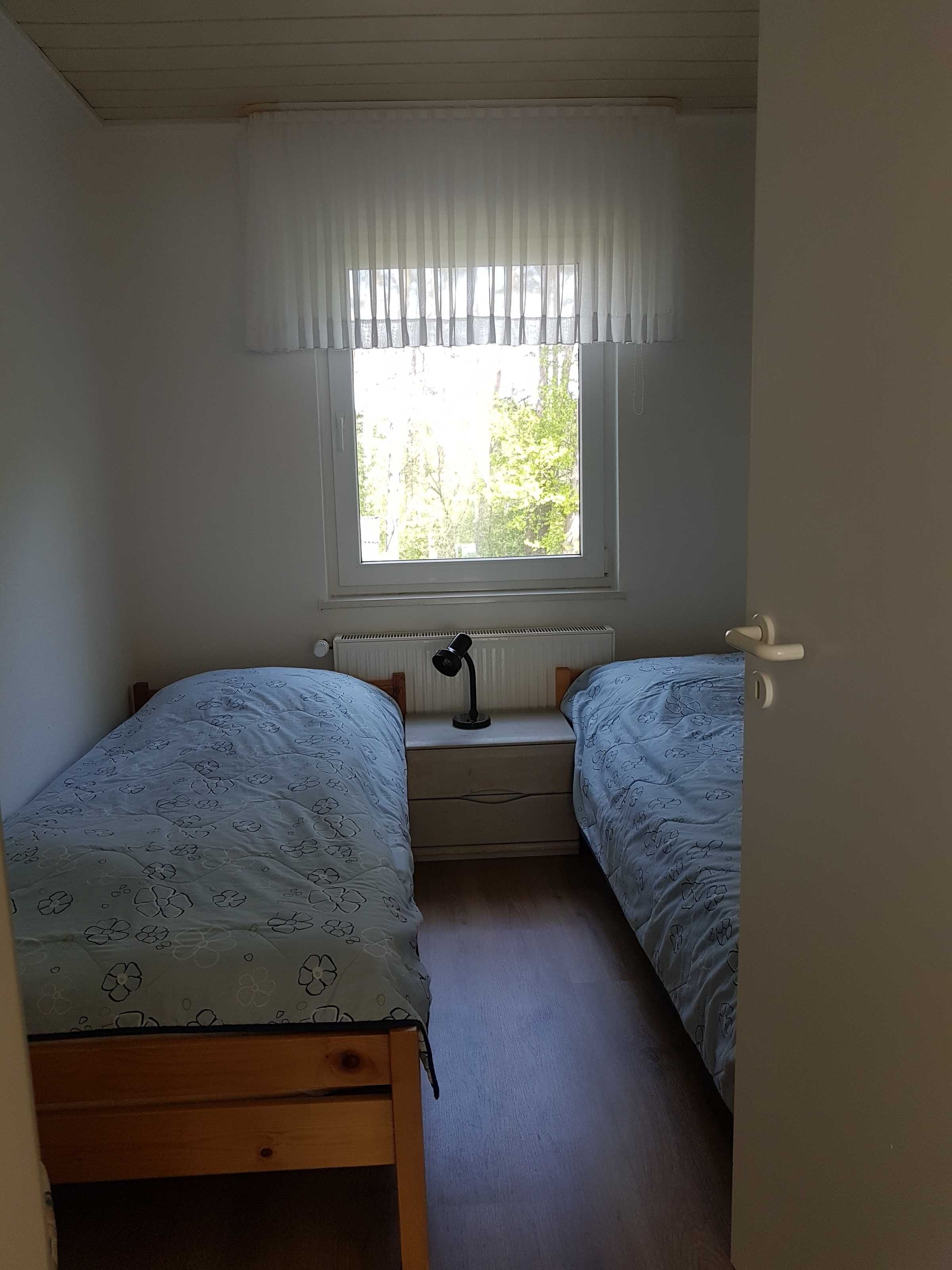 Een kleine slaapkamer met twee eenpersoonsbedden, een raam met witte gordijnen, een nachtkastje met een lamp en een radiator.