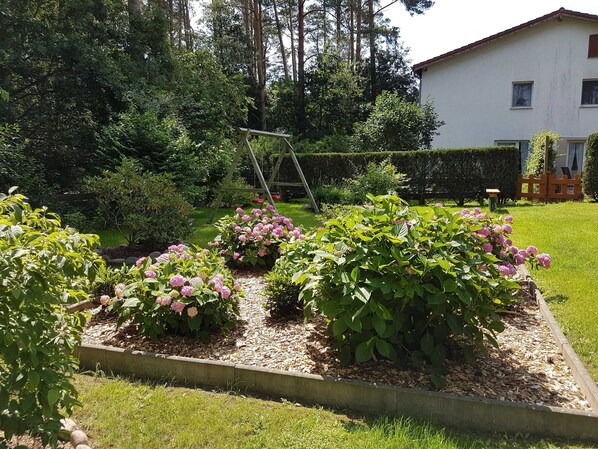 Ein Garten mit erhöhten Blumenbeeten, einer Holzliege und einem weißen Gebäude im Hintergrund.