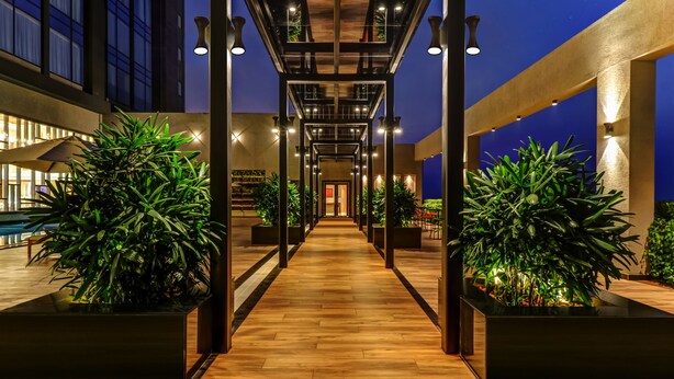 Un pasillo moderno de hotel con suelo de madera, grandes macetas y un pasillo central flanqueado por columnas.