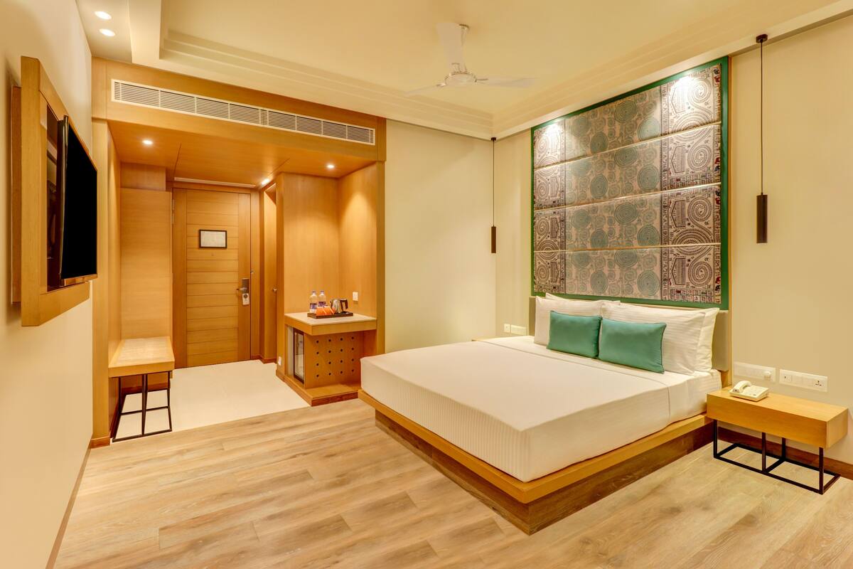 A modern hotel room with a large bed, a bedside table, a small desk, a wardrobe, and a television.