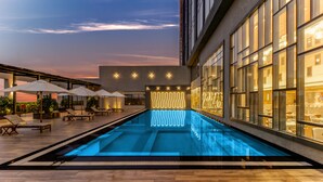 Una piscina con sillas de descanso y sombrillas, rodeada por un edificio de grandes ventanales y diseño moderno.