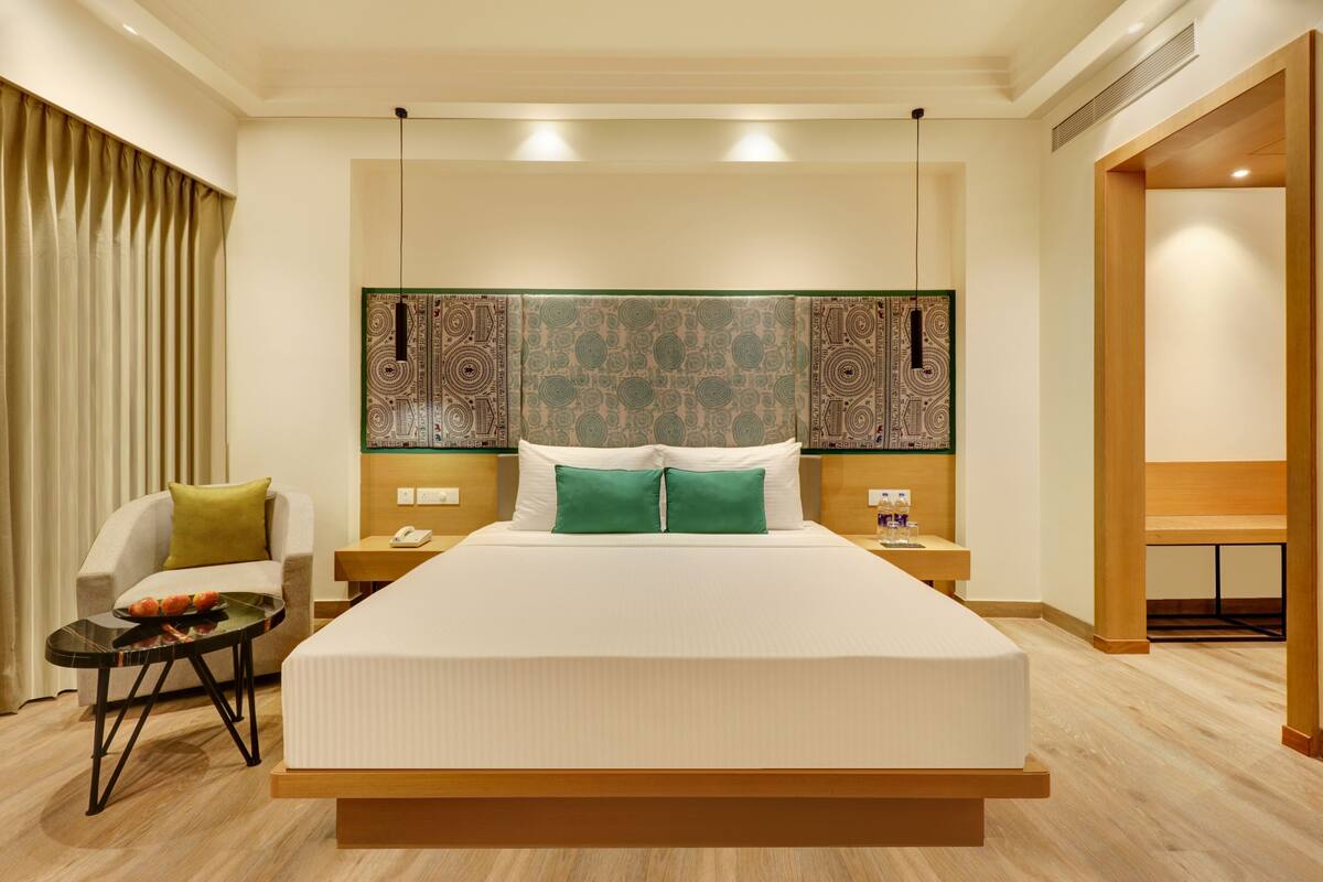 A modern hotel room with a large bed, a bedside table with a phone, a chair with a green pillow, and a decorative headboard.