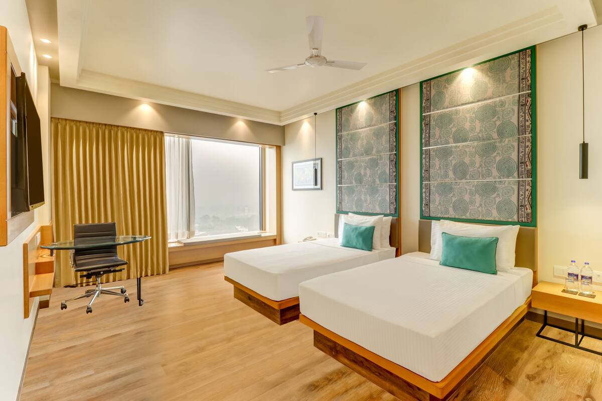A hotel room with two beds, a desk, a chair, a window with curtains, and a wall-mounted TV.