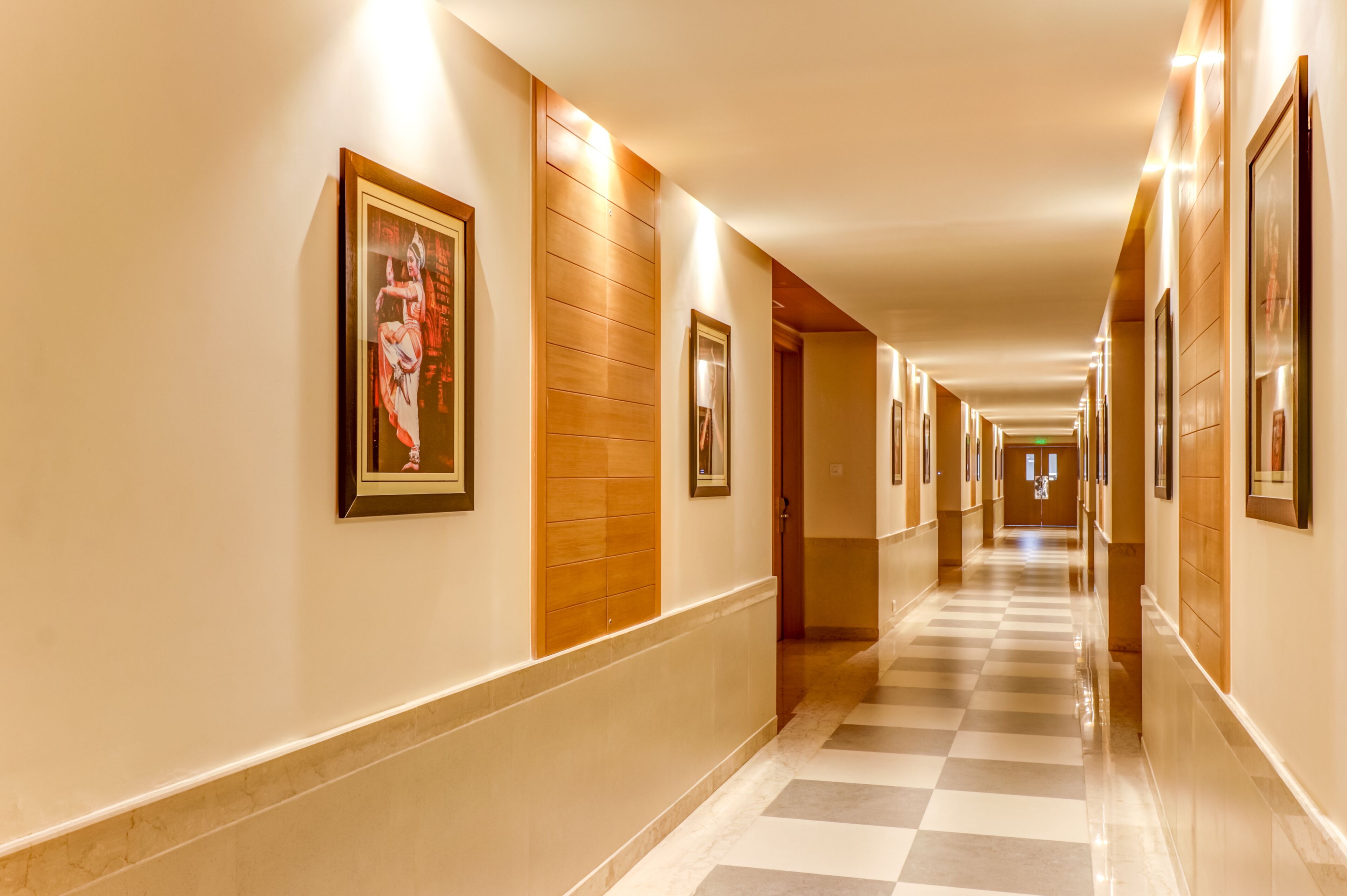 A hotel hallway with framed artwork on the walls, a checkered floor, and a wooden panel accent.
