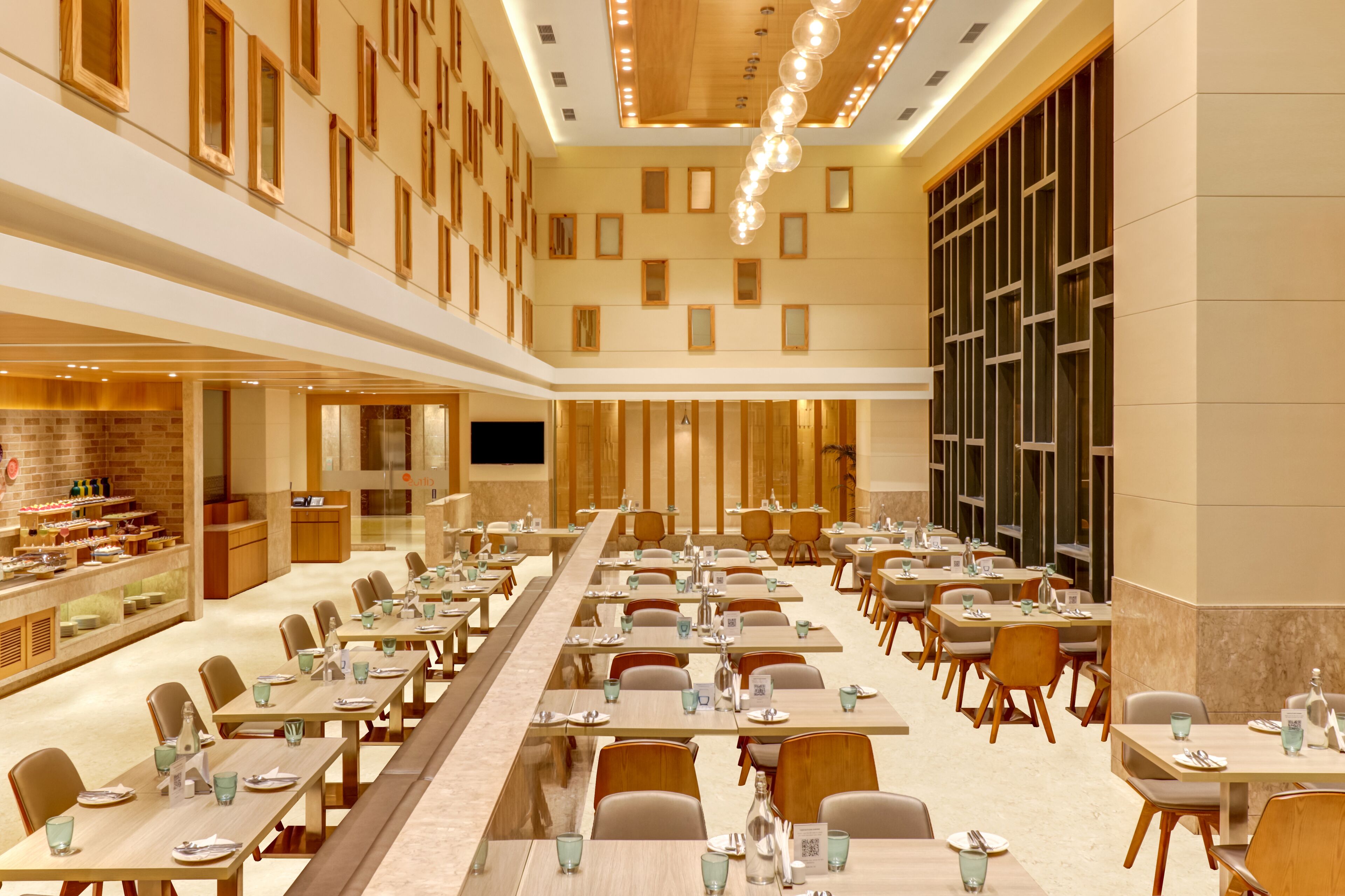 A spacious hotel dining area with multiple tables and chairs, a buffet on the left, and a large screen on the right.