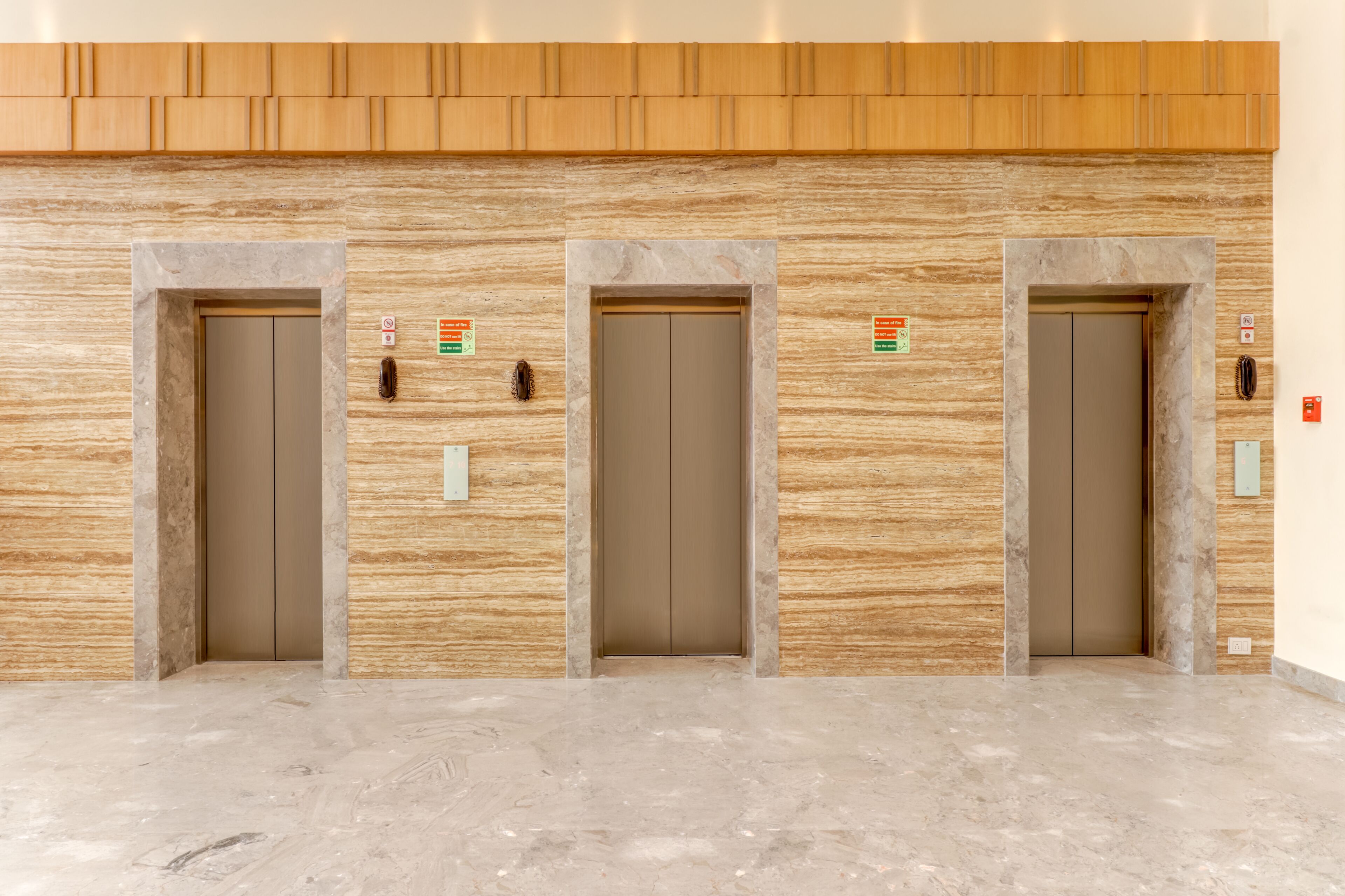 Three elevator doors in a lobby with a wooden panel wall and marble flooring.