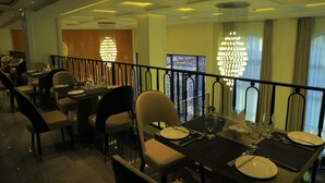 Une salle à manger avec des tables dressées pour quatre personnes, chacune agrémentée d’assiettes, de couverts et de verres. Un lustre est suspendu au plafond, et la pièce présente un design moderne, avec un balcon donnant sur un jardin.