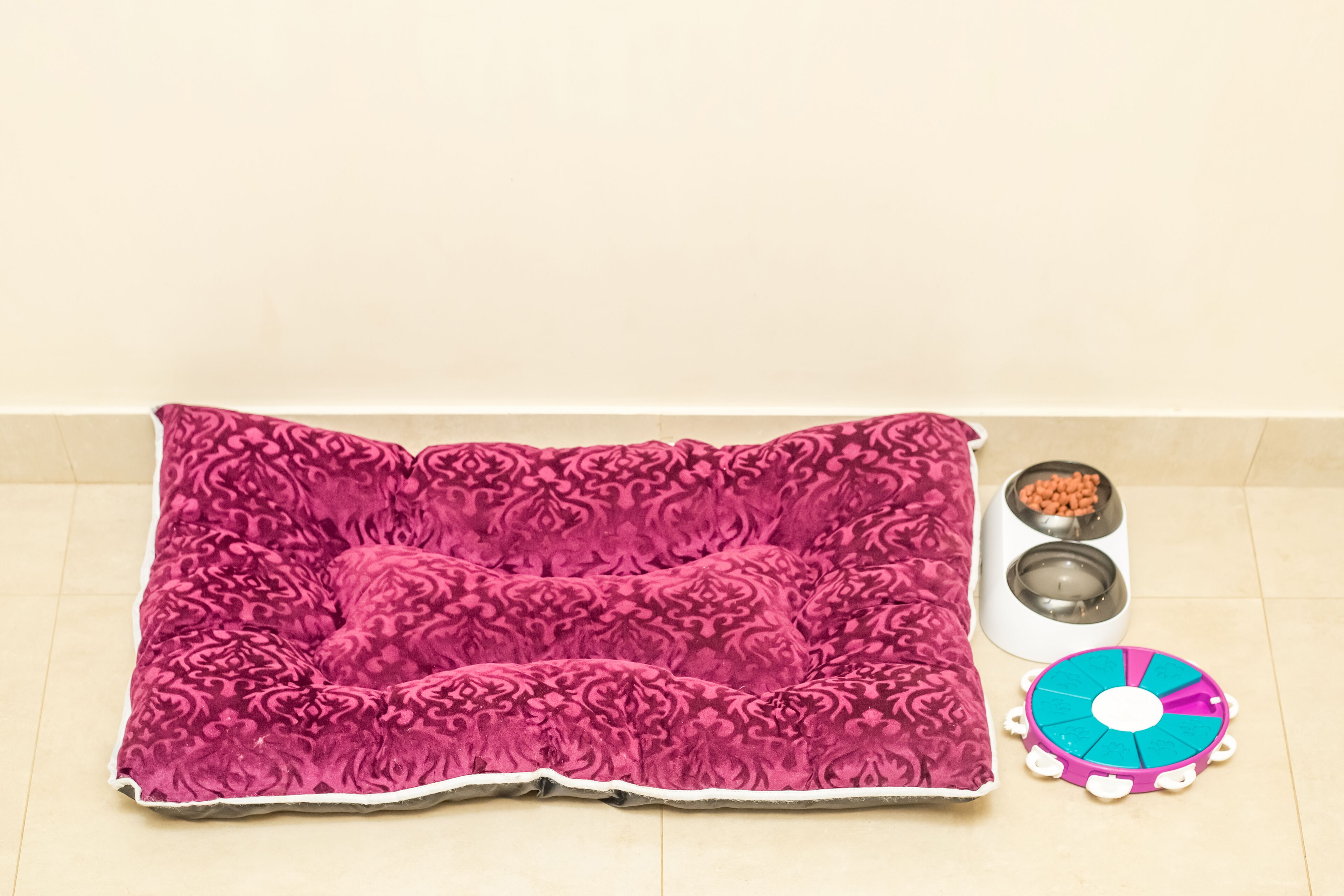 Una cama para mascotas con funda estampada, dos cuencos de comida para animales y un juguete rompecabezas sobre un suelo de baldosas.