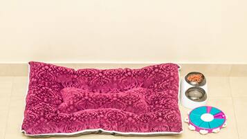 Una cama para mascotas con funda estampada, dos cuencos de comida para animales y un juguete rompecabezas sobre un suelo de baldosas.