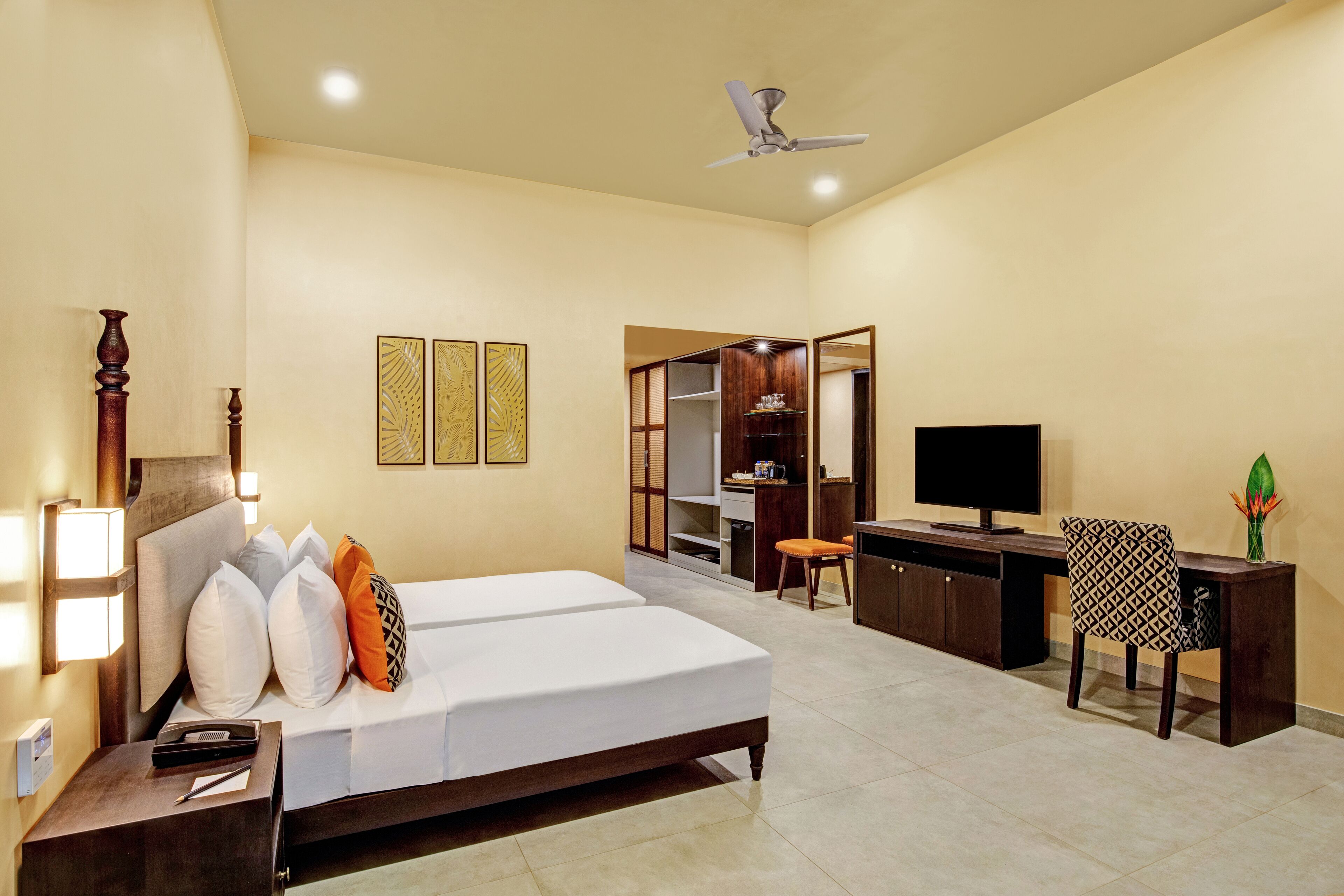 Habitación de hotel con cama, mesita de noche, lámpara, armario, escritorio, silla, televisor y ventilador de techo.