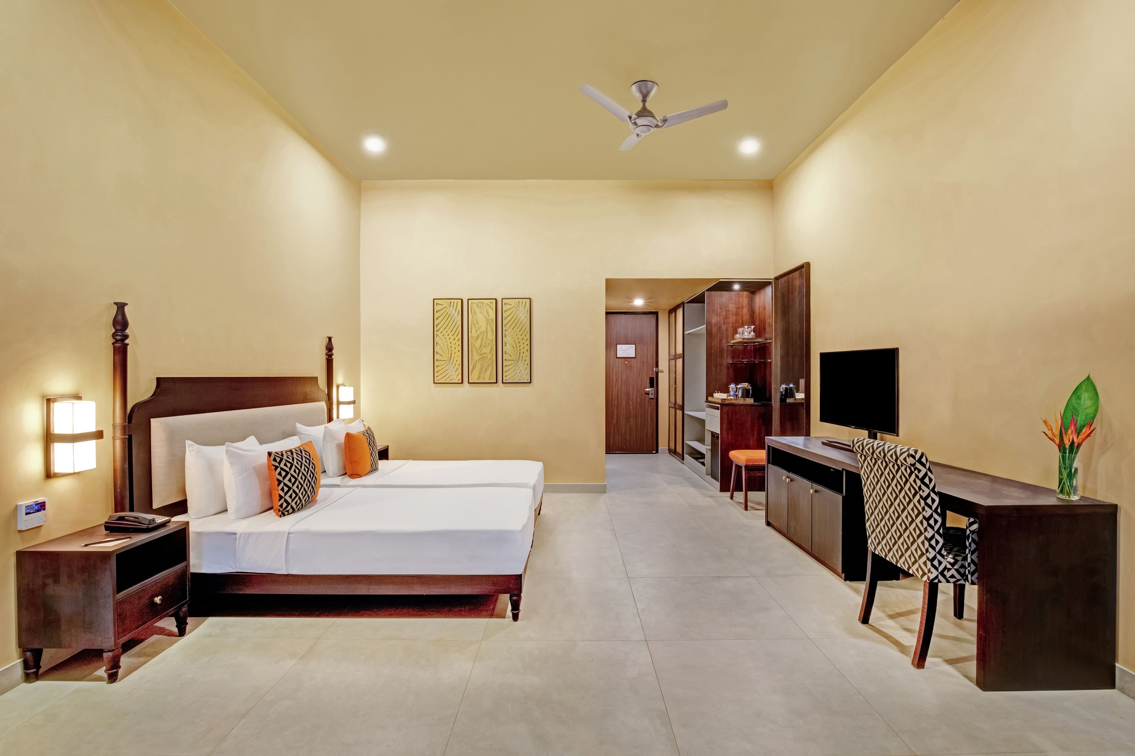 A hotel room with a bed, bedside table, lamp, desk, chair, TV, wardrobe, and ceiling fan.