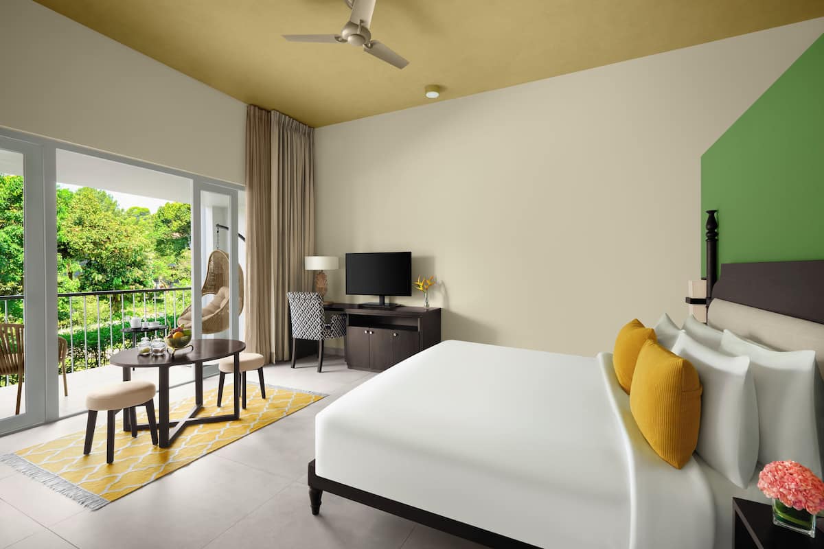 A hotel room with a bed, a desk with a TV, a chair, and a balcony with a view of greenery.