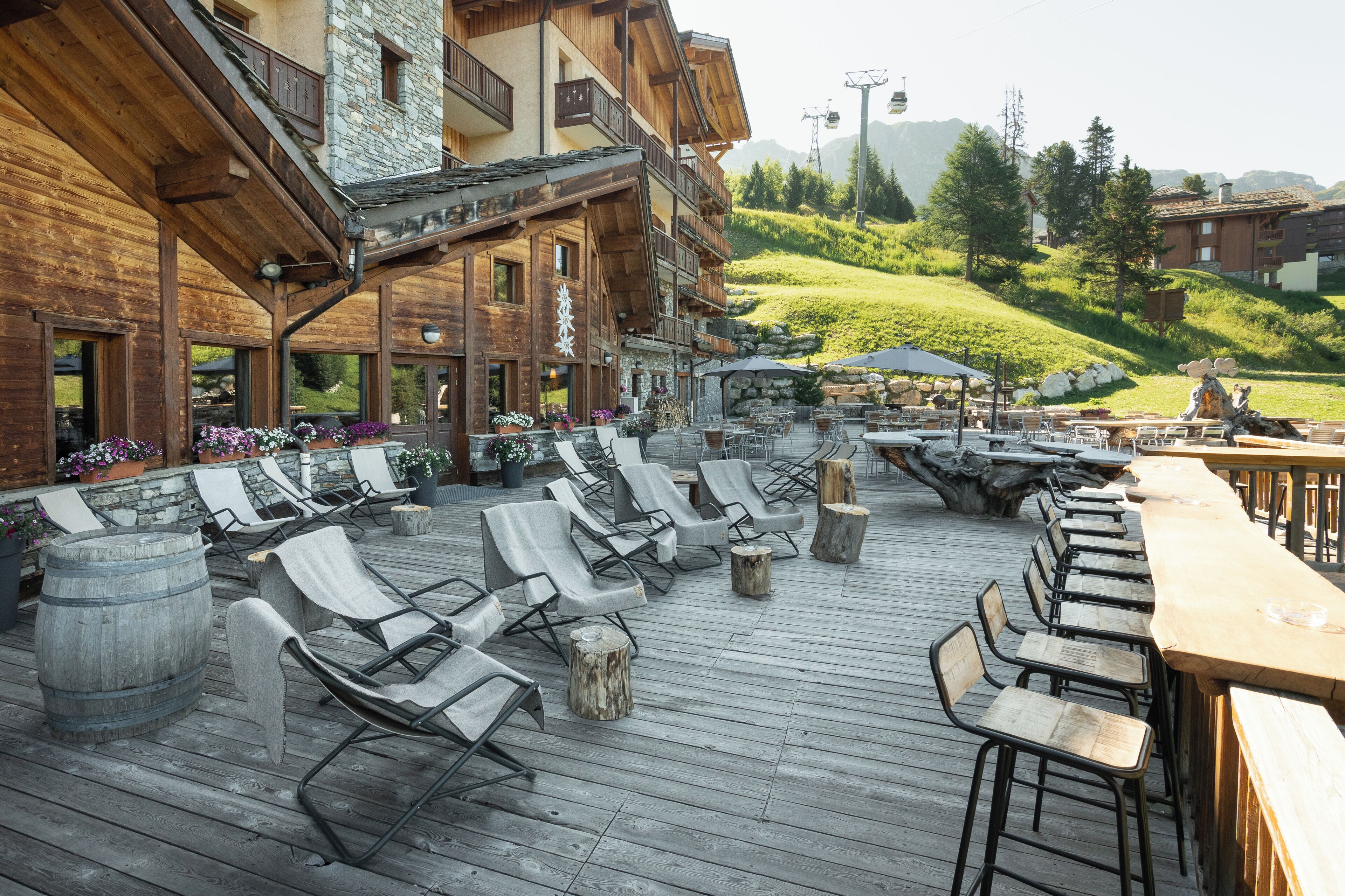 A wooden deck with lounge chairs and tables, a stone wall, and a view of a green hillside.