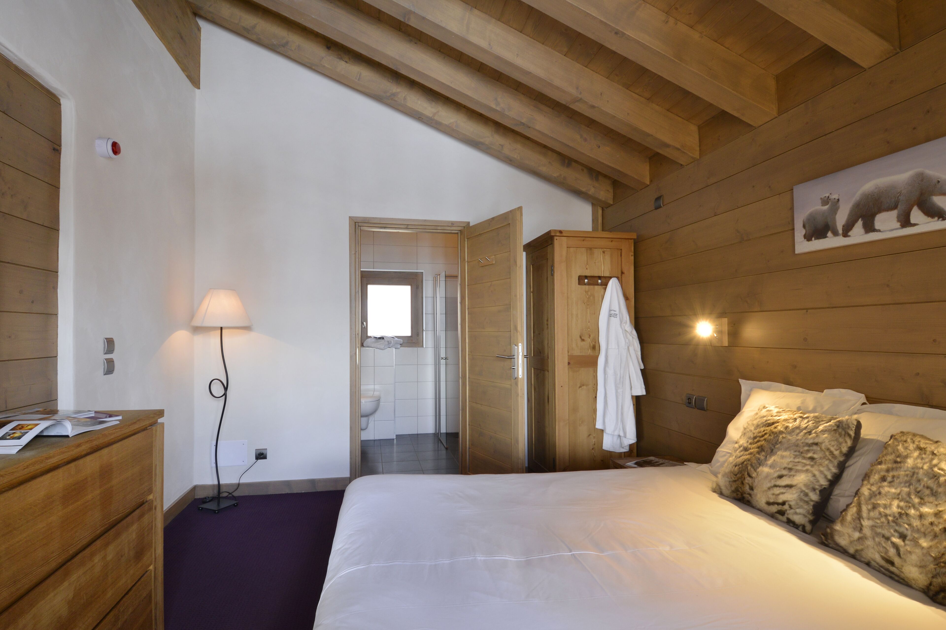 A bedroom with a wooden ceiling, a bed with white bedding, a wooden dresser, a lamp, and a polar bear painting on the wall.