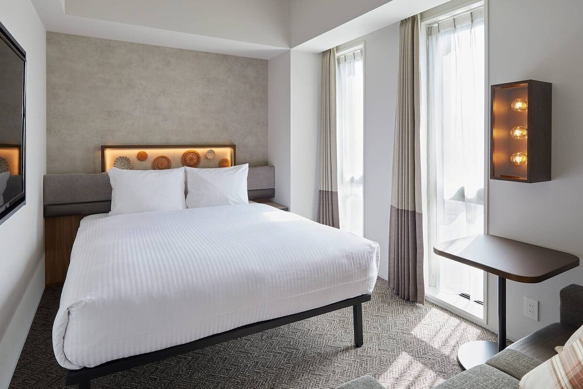 Hotellihuone, jossa on suuri sänky, yöpöytä, peili ja ikkuna, jossa on verhot.
