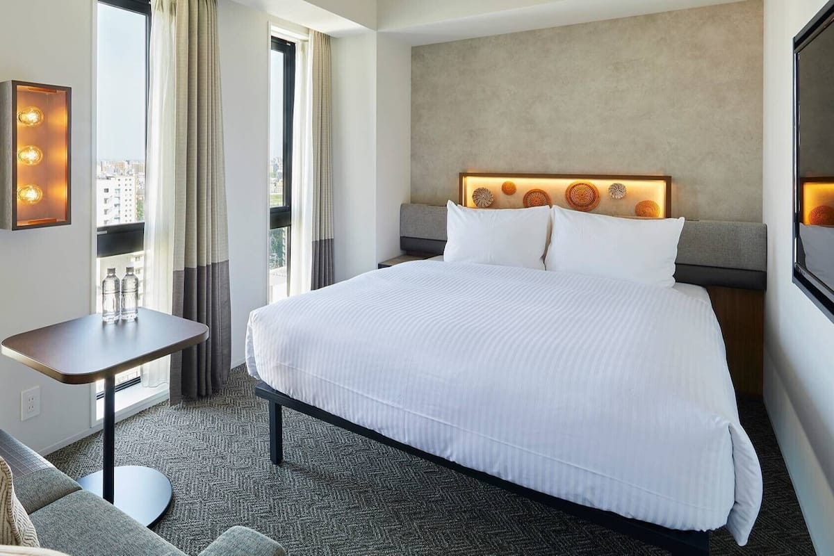 Hotellihuone, jossa on suuri sänky, pieni pöytä, sohva ja ikkunasta avautuva näkymä kaupunkiin.