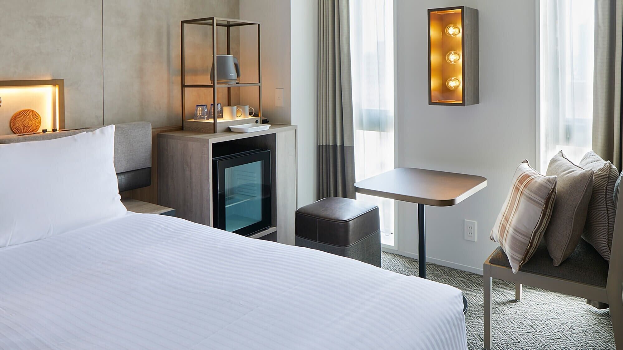 Habitación de hotel con cama, una mesita, zona de estar, minibar y una lámpara fijada a la pared.