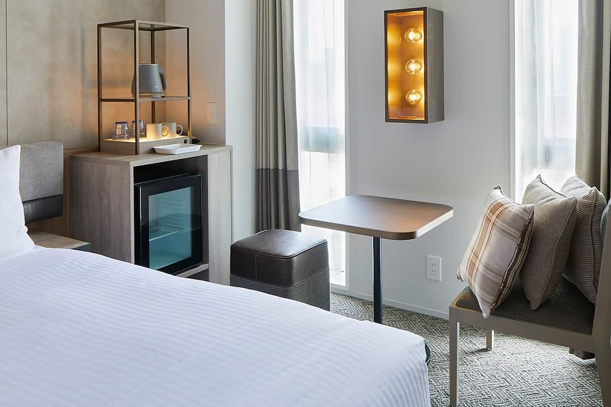 Hotellihuone, jossa on sänky, pieni pöytä, oleskelualue, minibaari ja seinään kiinnitetty valaisin.
