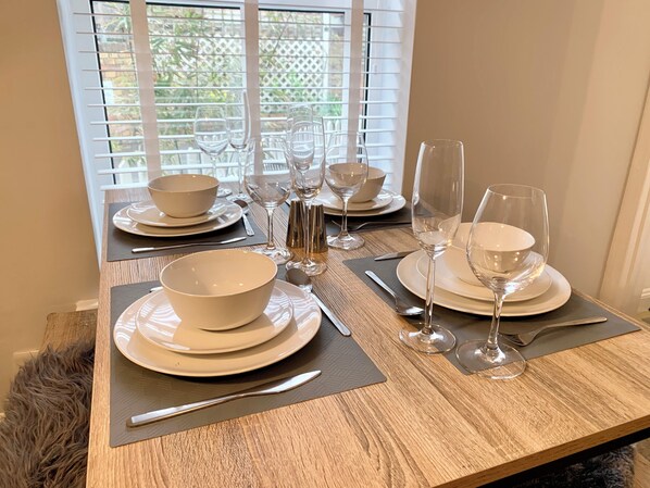 A dining table set with white plates, bowls, and wine glasses, with a view of greenery through the window.