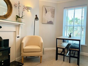 A room with a beige armchair, a small table with a chair, a floor lamp, a wall-mounted mirror, and a window with blinds.