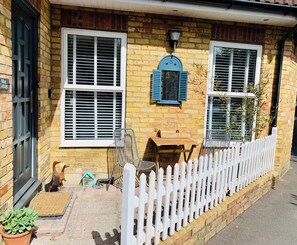 A brick house with two windows, a white picket fence, a wooden table, and a chair outside.