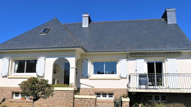 Une maison à deux étages, avec des fondations en pierre, une grande véranda à l’avant et un balcon doté d’une balustrade.