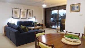 A living room with a dark blue sofa, yellow chairs, a wooden dining table, and framed pictures on the wall. - Braemar Unit 3 (Forster)