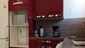 Eine kompakte Küche mit roten Schränken, einem Edelstahlkühlschrank, einer Kaffeemaschine und einer Mikrowelle.