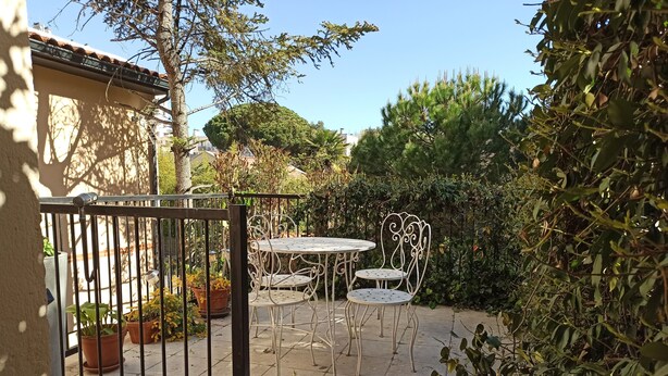 Eine Terrassengruppe im Freien mit rundem Tisch und zwei Stühlen, umgeben von Kübelpflanzen und einem Metallzaun.